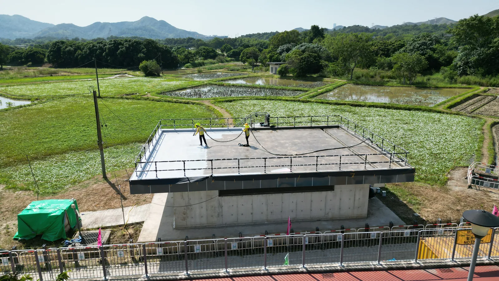 古洞機房天台的高角度視圖,兩名工人在田野環繞的屋面施工