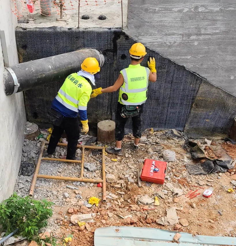 Two workers positioning black textured sheeting beside a wall base and large pipe at No. 3 Middle Gap Road