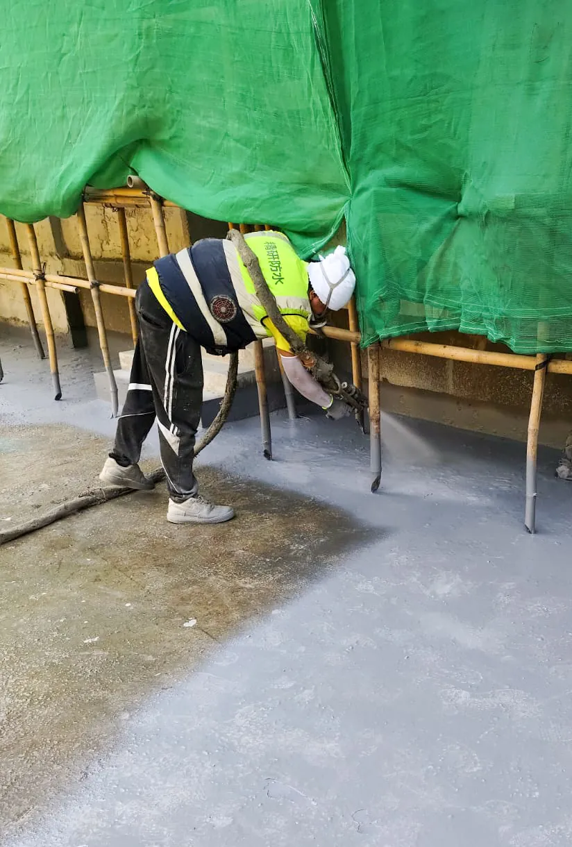 Worker spray-applying a grey surface along a ground edge beneath green scaffold netting at No. 3 Middle Gap Road