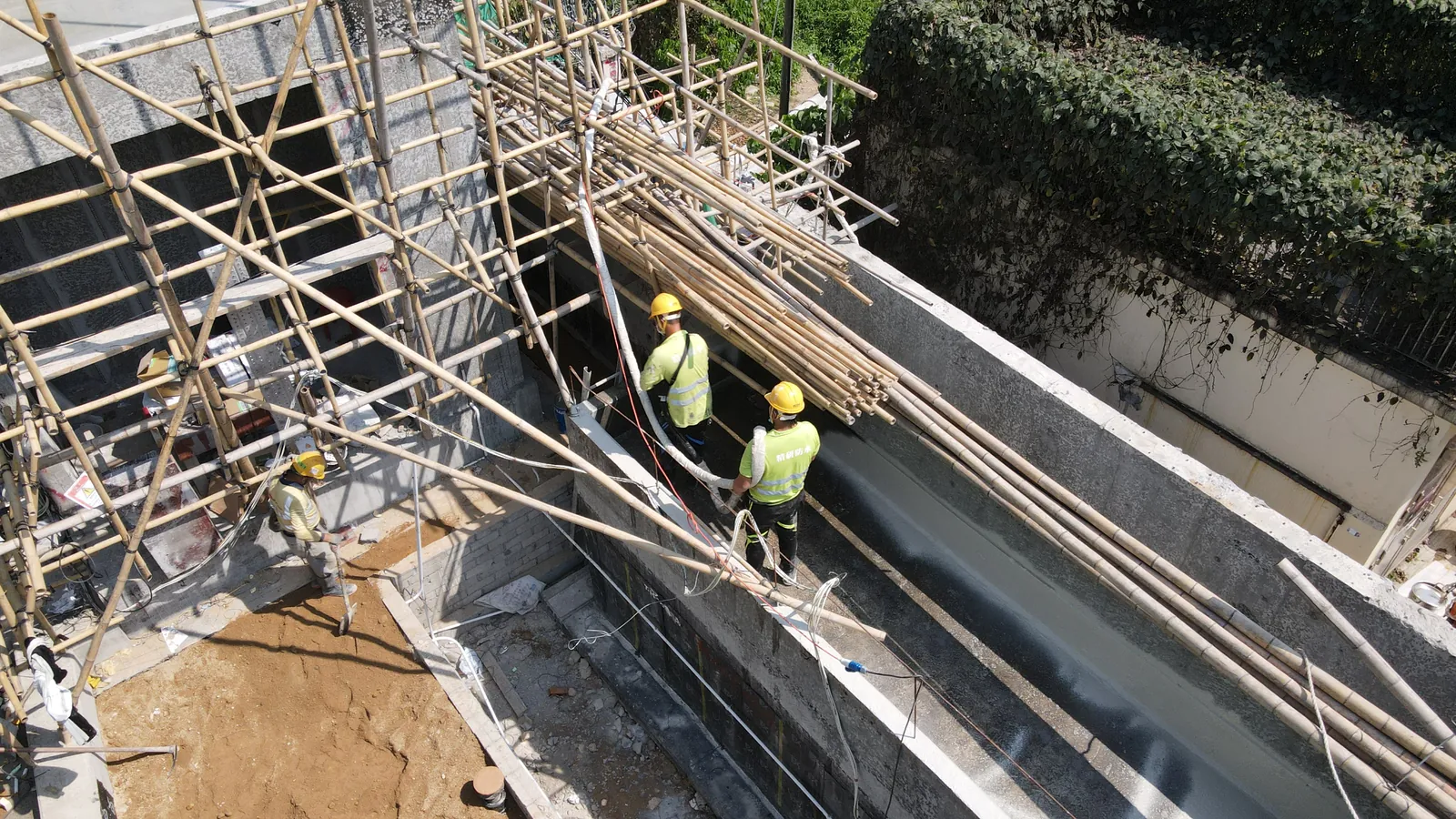 Three workers on bamboo scaffolding beside a long grey wall at the Villa Panova site in Sai Kung.