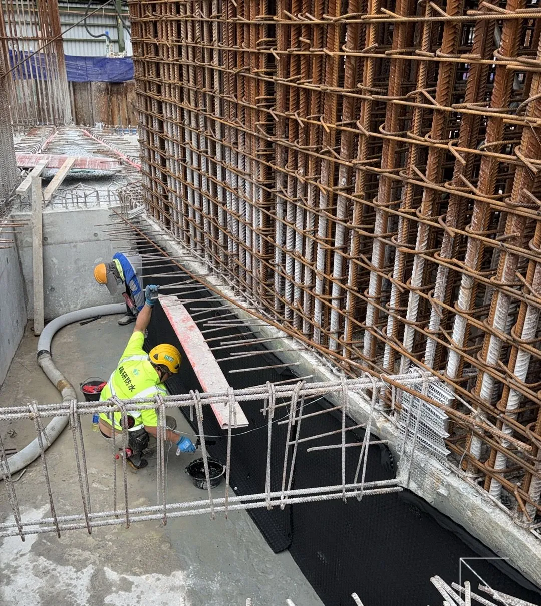 Workers applying dark material across the base of a pit beside reinforced walls at the Wang Chau public housing site